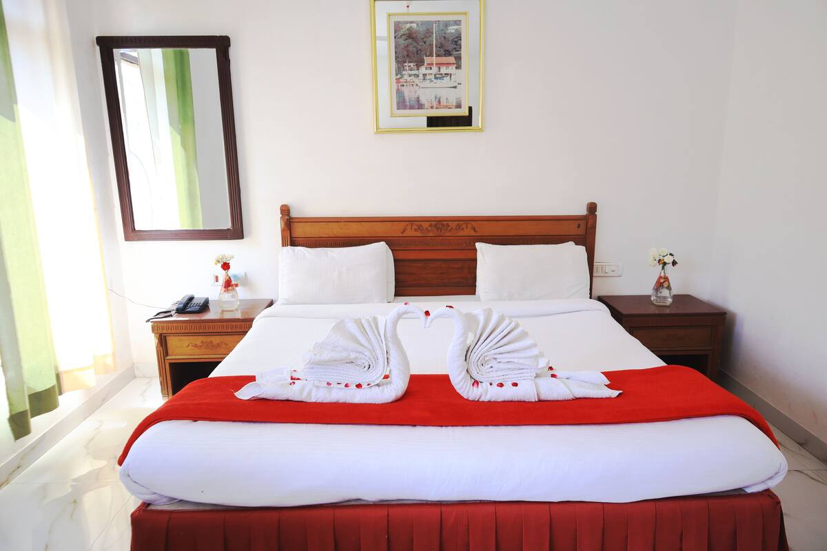 A neatly made bed with a red blanket and two swan-shaped towels, a wooden headboard, white pillows, a nightstand with a phone and a vase of flowers, and a framed picture on the wall.