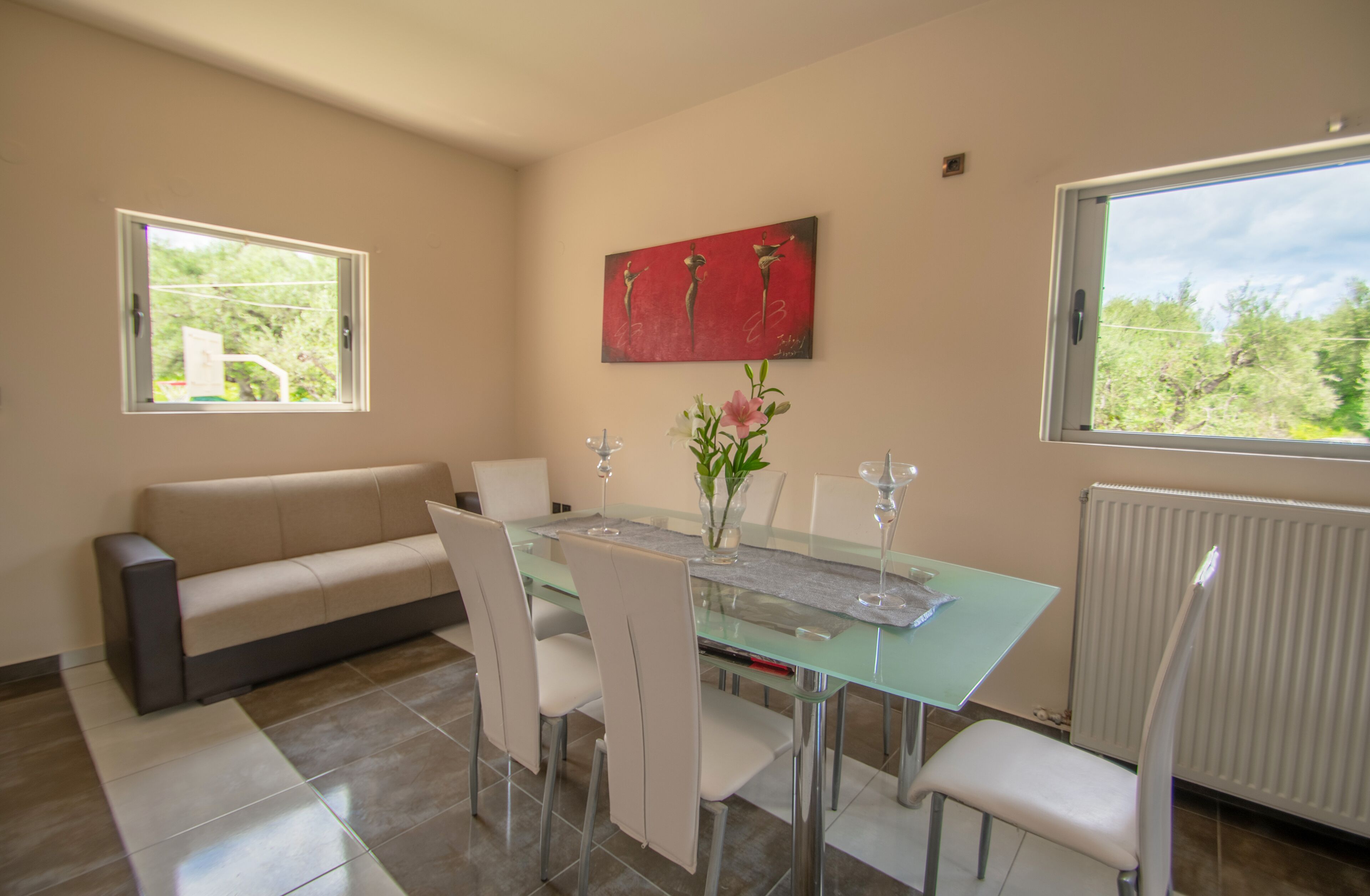 Uma área de refeições com mesa de vidro, cadeiras brancas, um sofá bege e um quadro vermelho na parede.