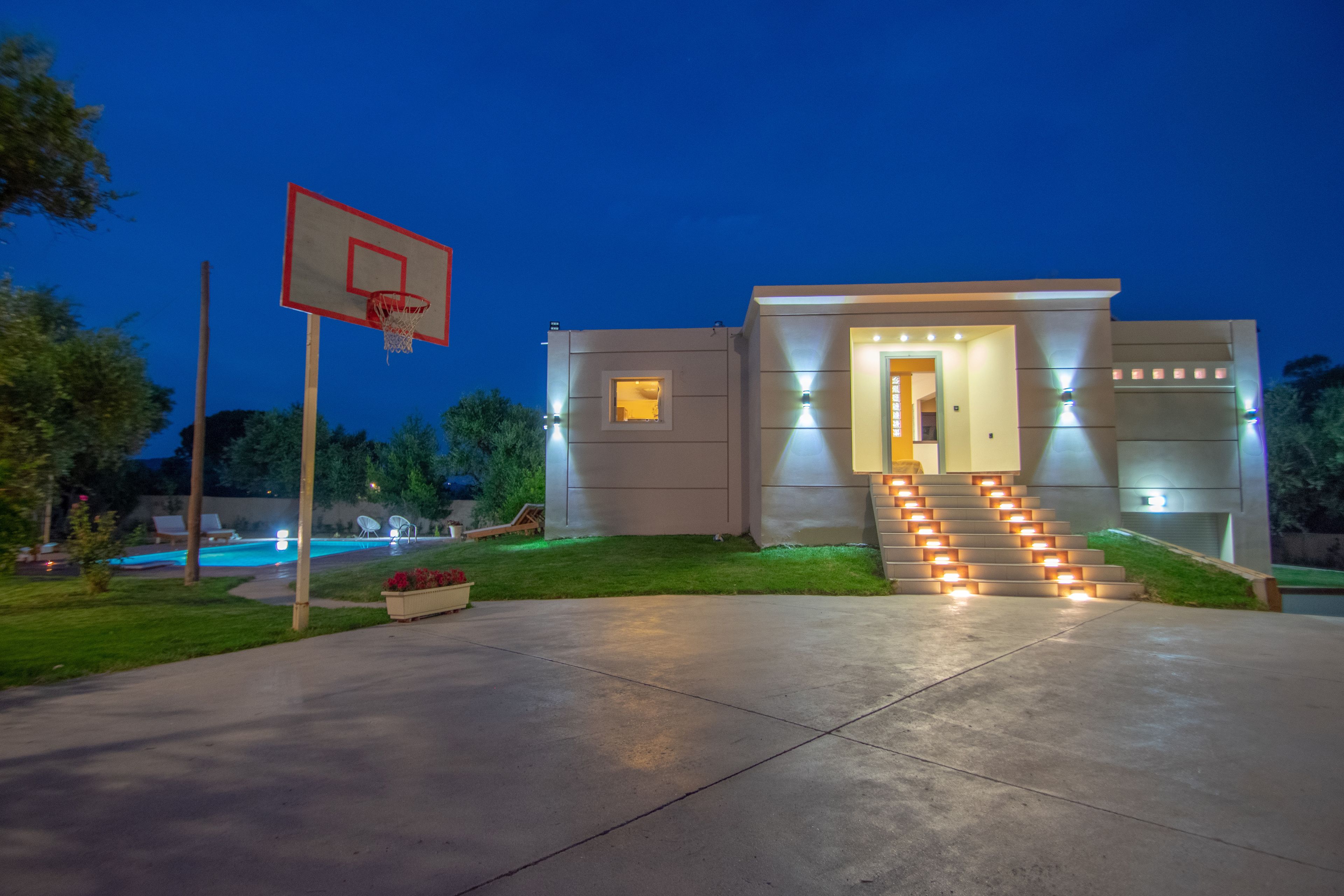 A modern house with a basketball court, a pool, and a well-lit entrance.