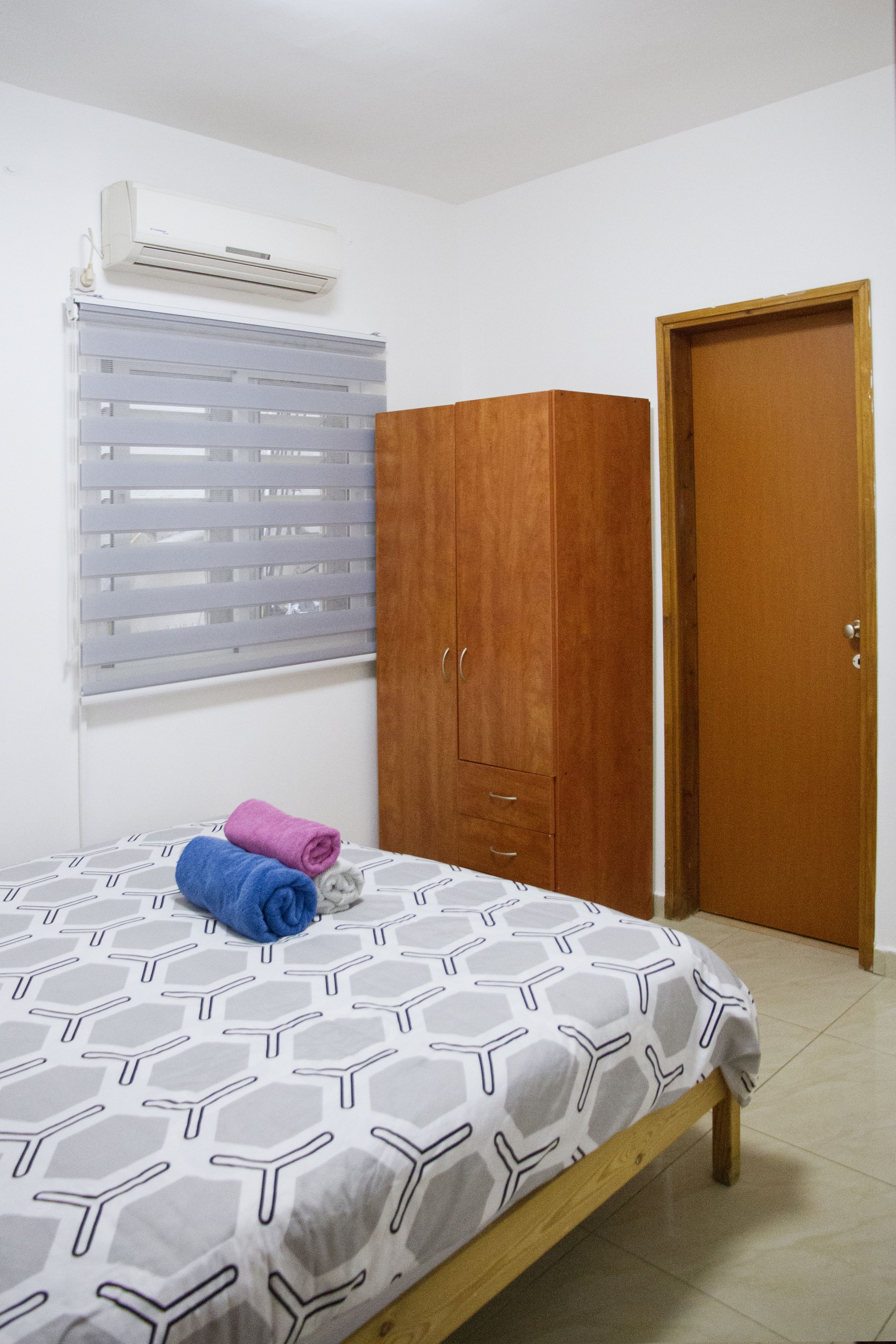 A bedroom with a bed, a wooden wardrobe, a window with blinds, and a door.