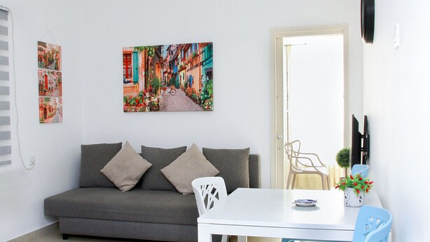 Ein Wohnzimmer mit einer Couch, einem Esstisch und Stühlen. Zwei Bilder hängen an der Wand, und eine Tür führt in einen anderen Raum.