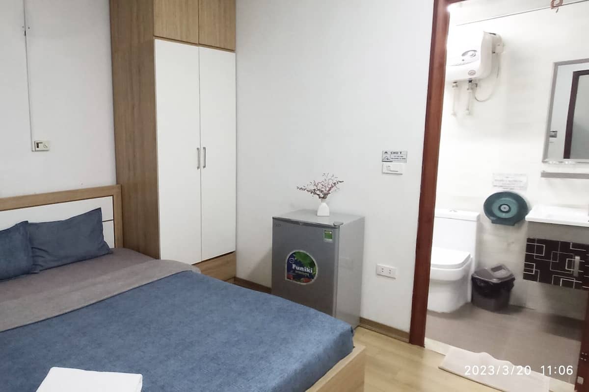 A bedroom with a bed, a nightstand, a small refrigerator, a plant, and a bathroom visible through an open door.