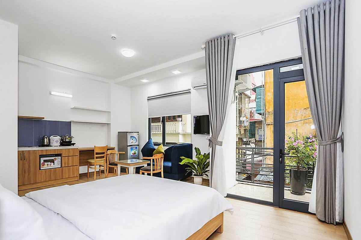 A modern hotel room with a bed, a small kitchenette, a dining area, and a balcony with curtains.