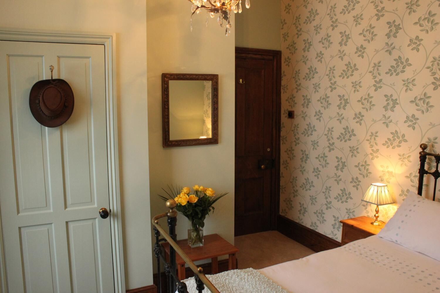 A bedroom with a bed, a nightstand with a lamp, a dresser, a mirror, and a door with a hat hanging on it.