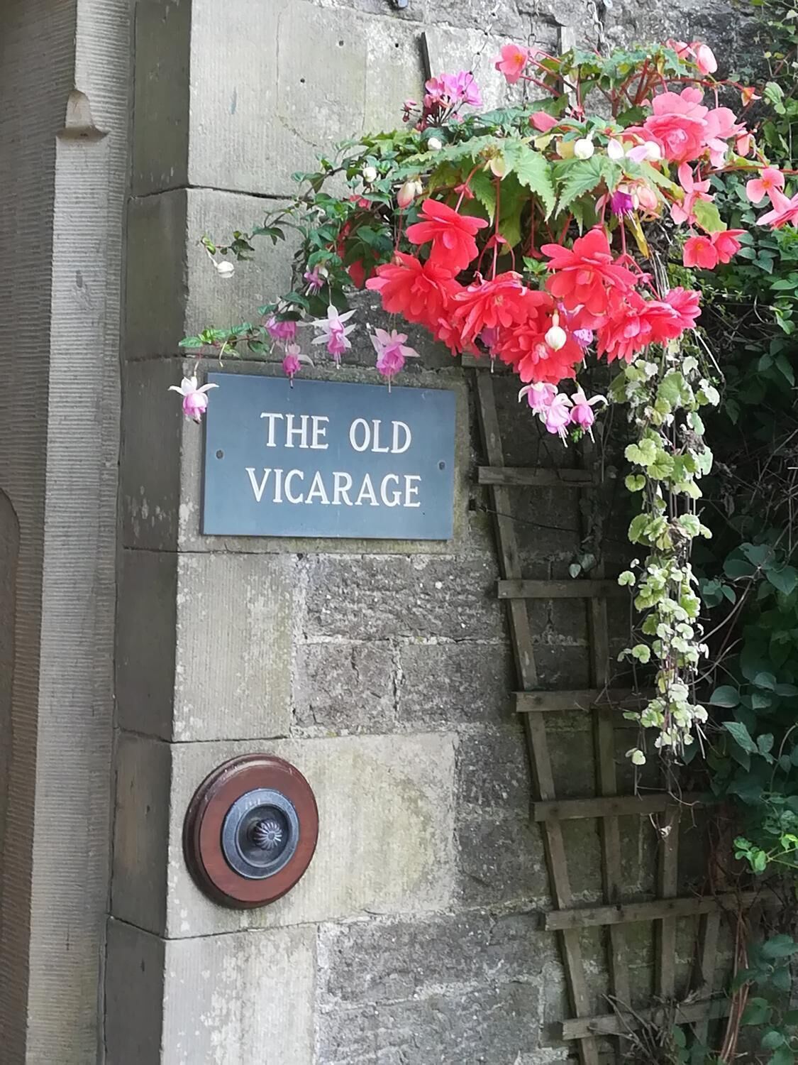 Une plaque portant l’inscription « THE OLD VICARAGE » est fixée à un mur de pierre, surmontée d’un treillage en bois et de plantes grimpantes en fleurs.