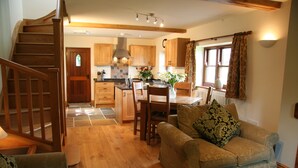 A cozy living room with a wooden staircase, a dining area with a table and chairs, and a kitchen with wooden cabinets and a window with curtains.