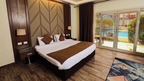 Une chambre d’hôtel avec un grand lit, une tête de lit en bois, une table de chevet, une lampe et une vue sur une piscine, visible à travers de grandes portes-fenêtres coulissantes.