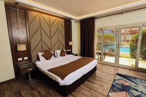 A hotel room with a large bed, a wooden headboard, a nightstand, a lamp, and a view of a pool through large sliding glass doors. - Byoot Bay Hotel & Resort (Imbabah)