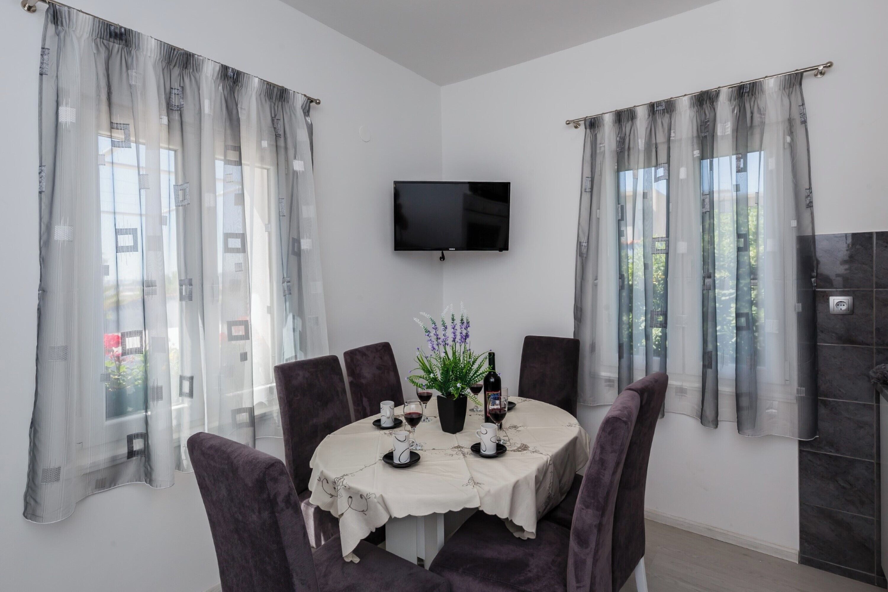 Un coin repas avec une table ronde, des chaises violettes, un téléviseur fixé au mur et des rideaux légers.