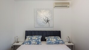 A bedroom with a bed, two nightstands, a wall-mounted air conditioner, and a floral painting. - Luxury villa complex Pendolino with heated pool and jacuzzi (Kastela)