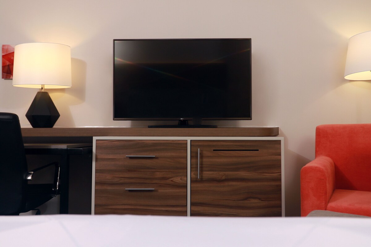 Une chambre d’hôtel équipée d’un téléviseur à écran plat fixé au mur, d’un bureau avec une lampe, d’une chaise et d’une commode avec des tiroirs.