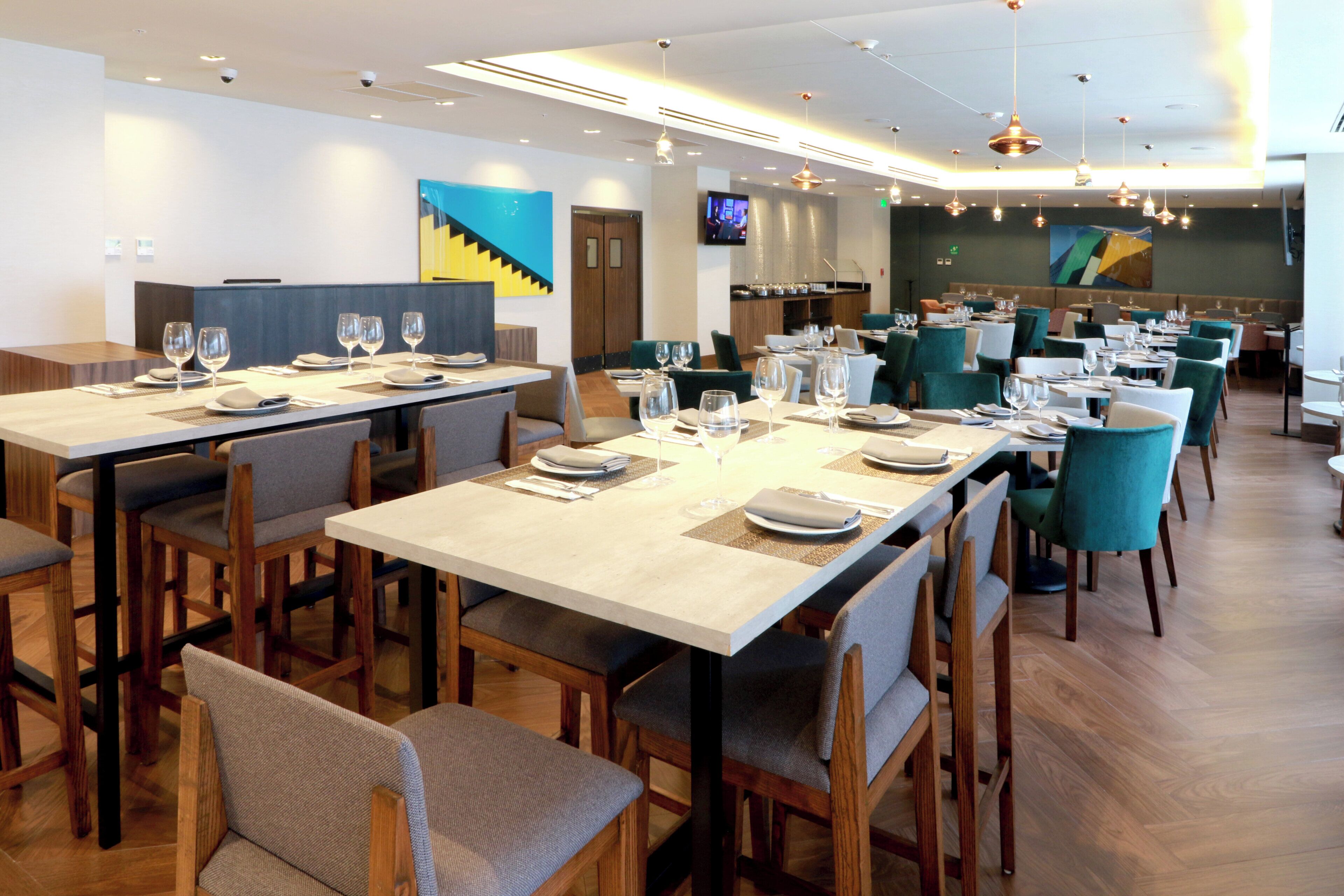 A modern restaurant with multiple dining tables and chairs, a flat-screen TV mounted on the wall, and a contemporary ceiling design.