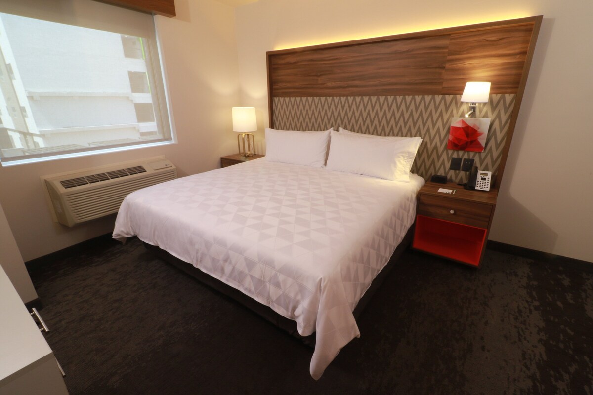 Une chambre d’hôtel avec un grand lit, une table de chevet avec une lampe, un meuble de rangement rouge, un téléphone fixé au mur et une fenêtre avec des stores.