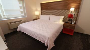 Quarto de hotel com cama grande, mesa de cabeceira com abajur, móvel de armazenamento vermelho, telefone fixado na parede e janela com persianas.