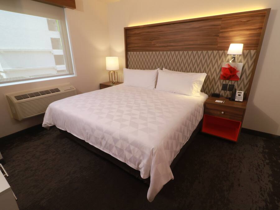 Quarto de hotel com cama grande, mesa de cabeceira com abajur, móvel de armazenamento vermelho, telefone fixado na parede e janela com persianas.