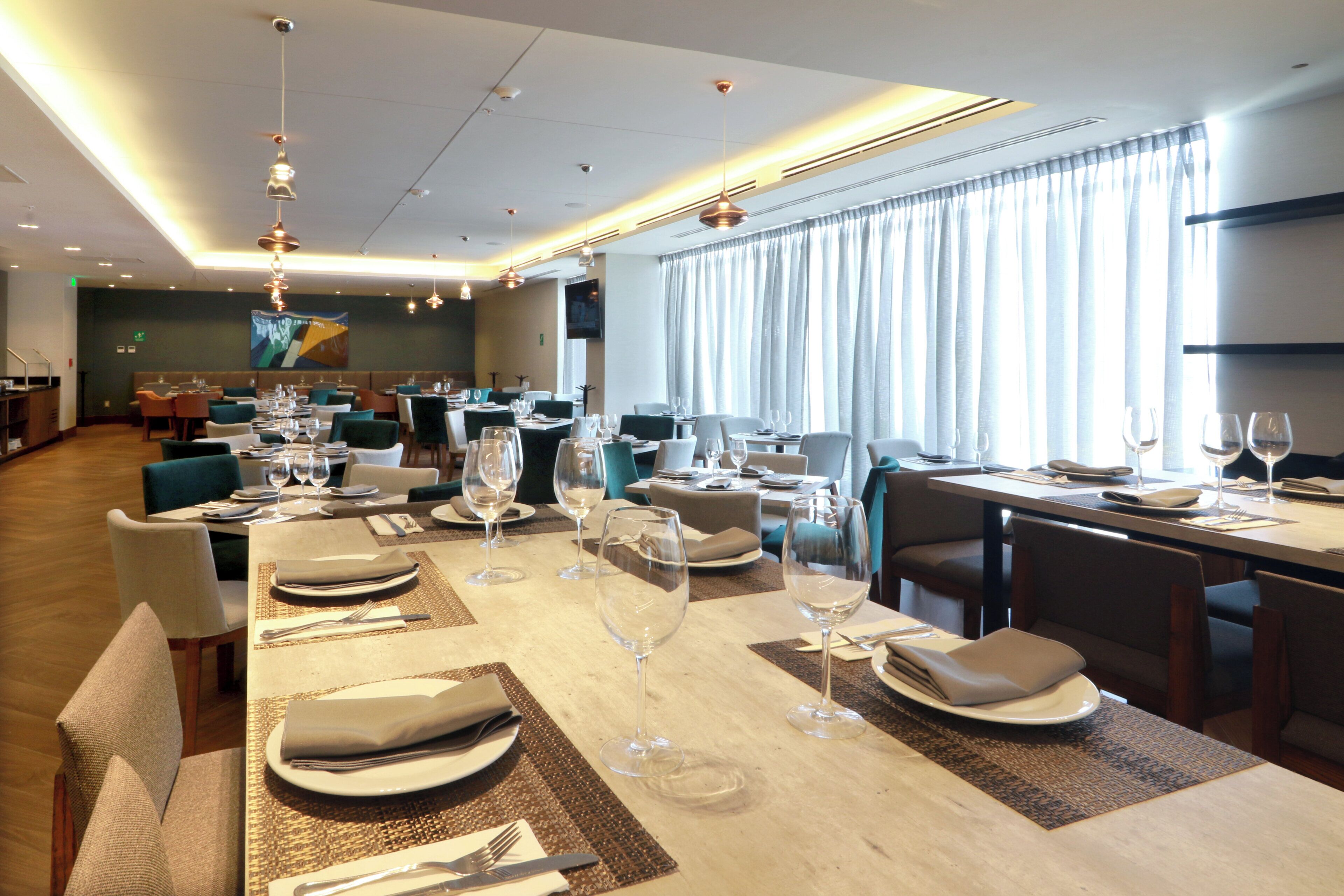A restaurant with multiple dining tables set with plates, glasses, and cutlery. There are hanging lights and a large window with curtains.