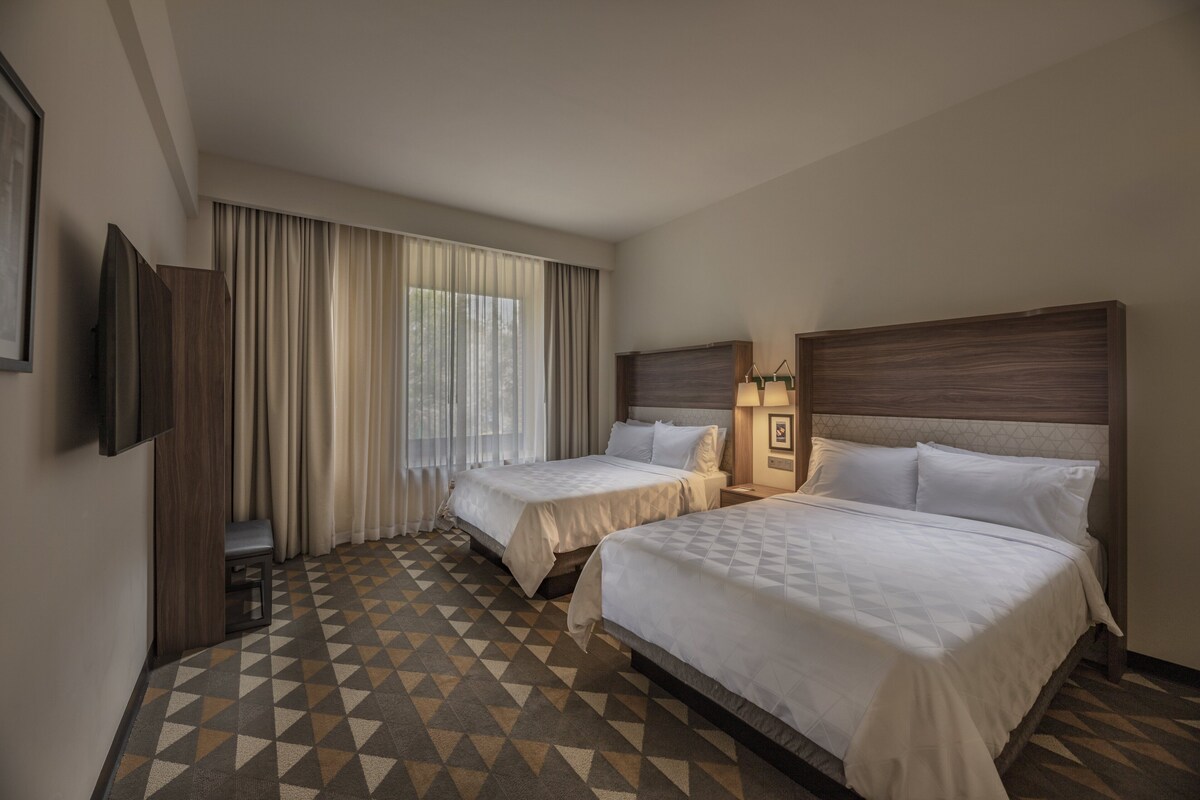 A hotel room with two beds, a TV, a small table, and a window with curtains.
