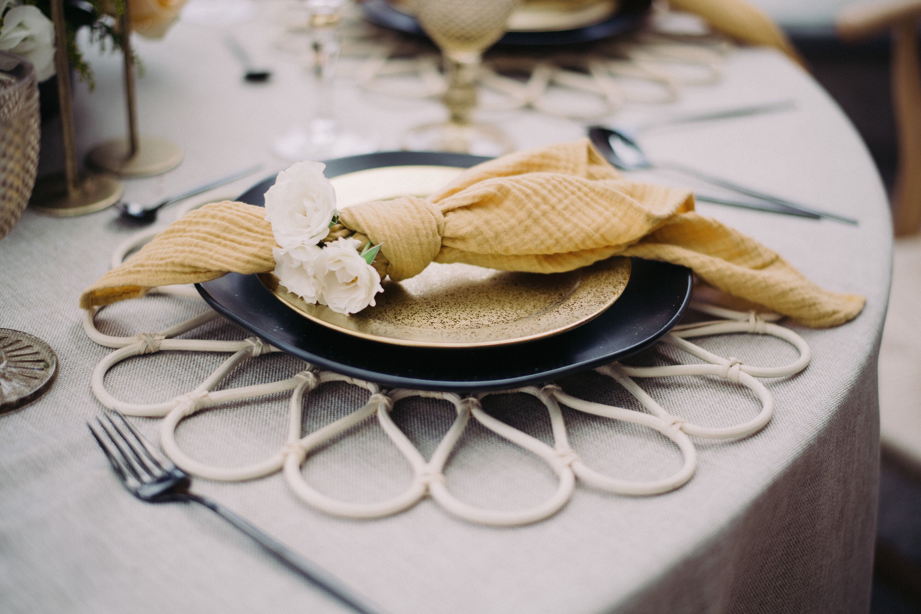 Una mesa dispuesta con un plato oscuro, una servilleta beige atada en lazo y un centro de mesa con una flor blanca.