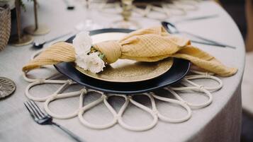 Una mesa dispuesta con un plato oscuro, una servilleta beige atada en lazo y un centro de mesa con una flor blanca.