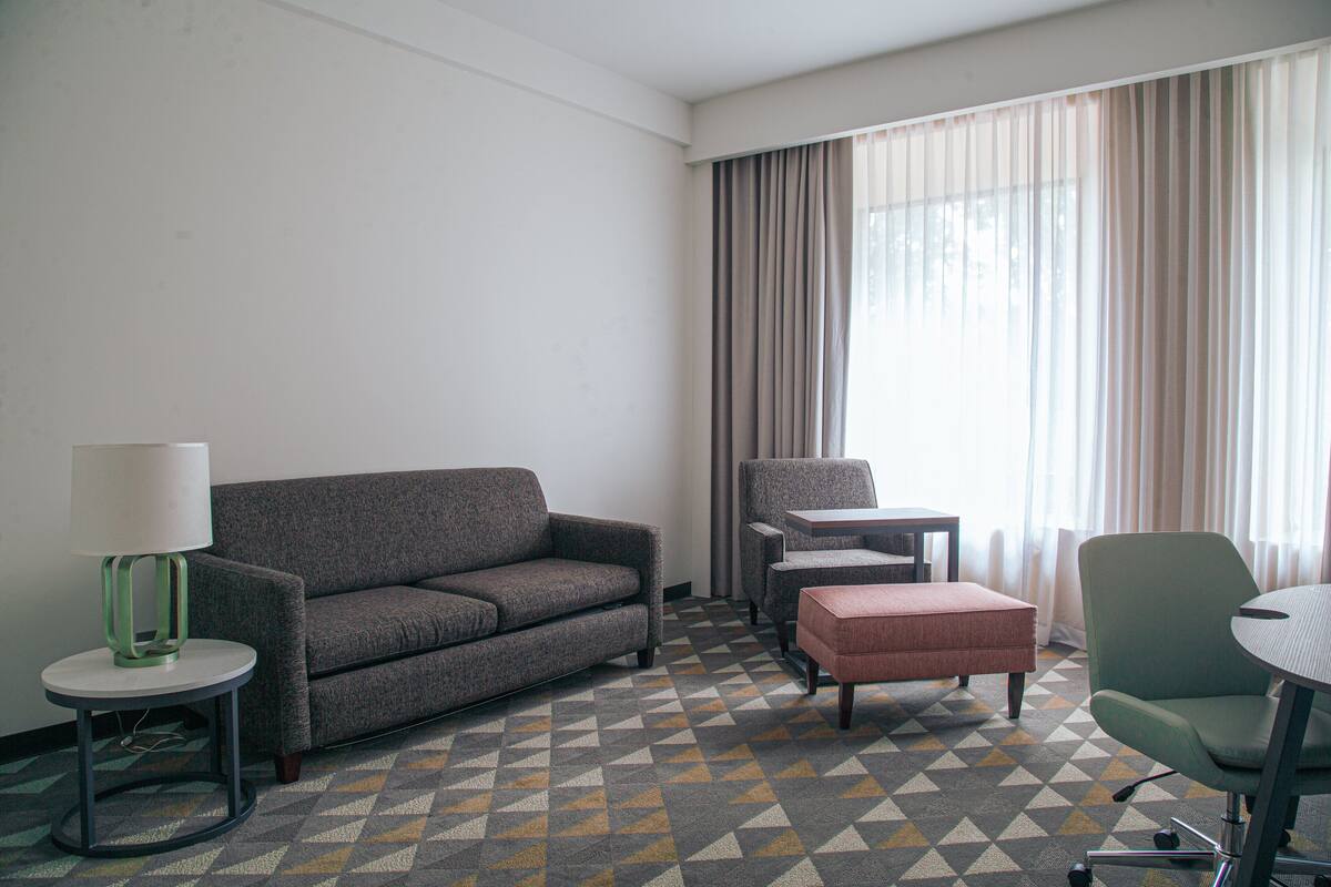 A hotel room with a gray sofa, a small round table with a lamp, a green armchair, and a patterned carpet.
