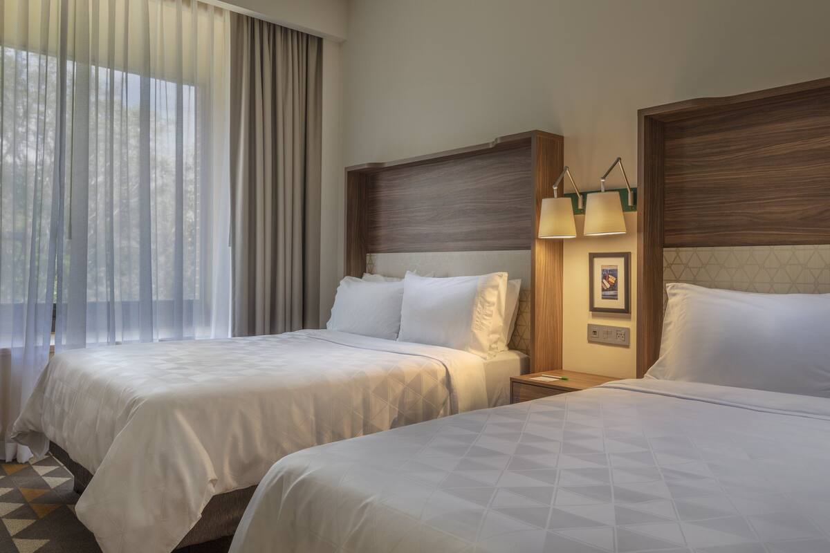 A hotel room with two beds, a nightstand with lamps, and a window with curtains.