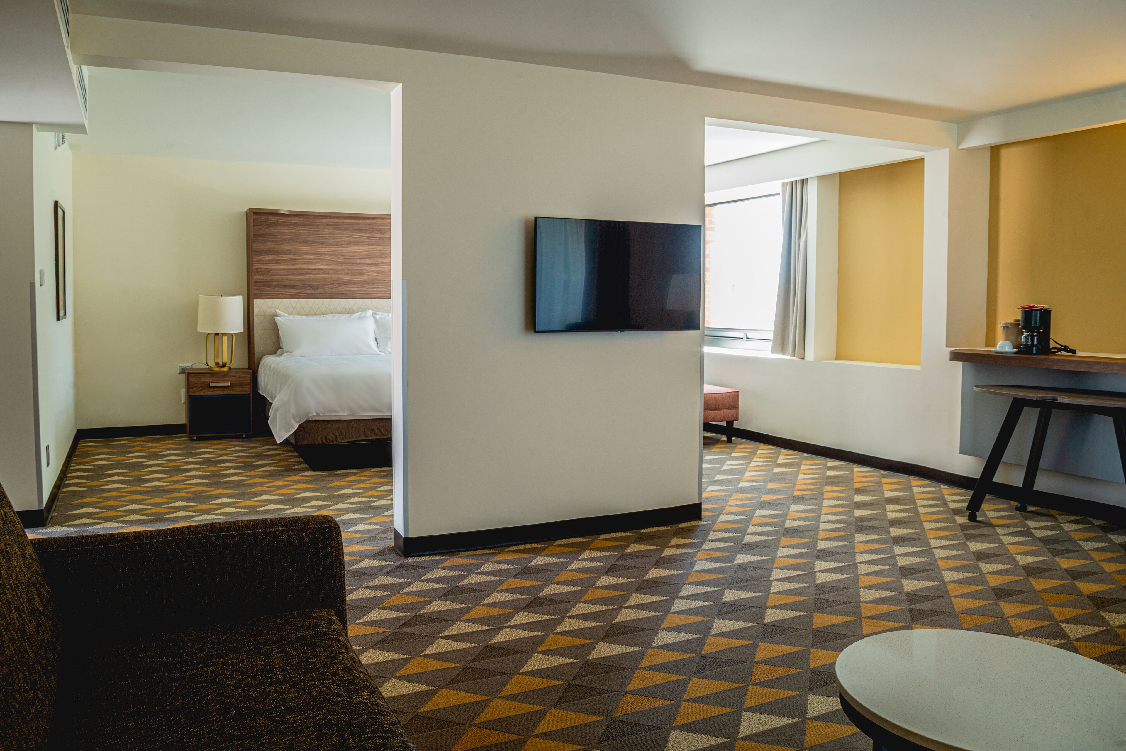 A hotel room with a bed, a TV mounted on the wall, a desk with a chair, and a patterned floor.