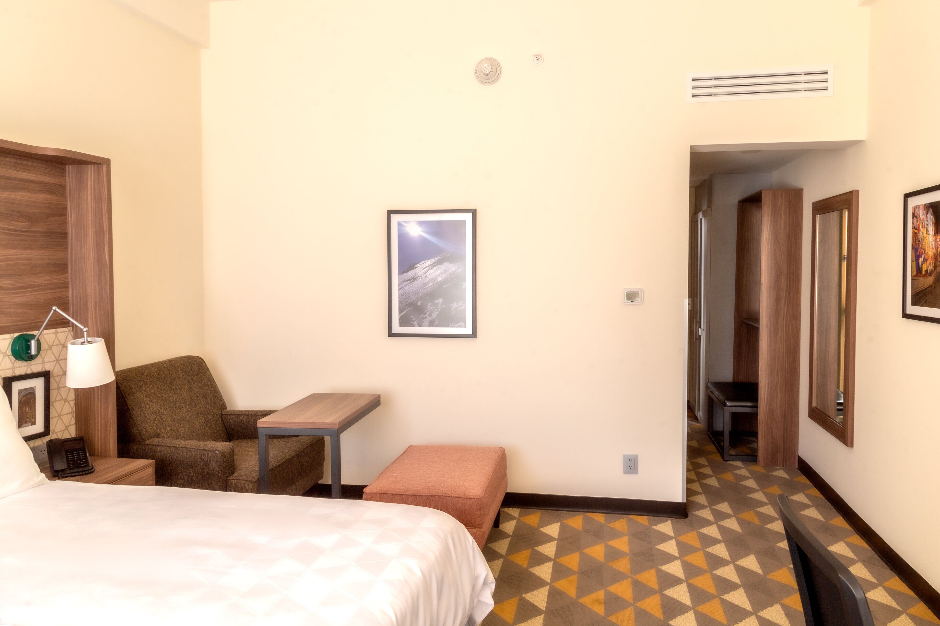 A hotel room with a bed, a desk, a chair, a small table, and a picture on the wall.