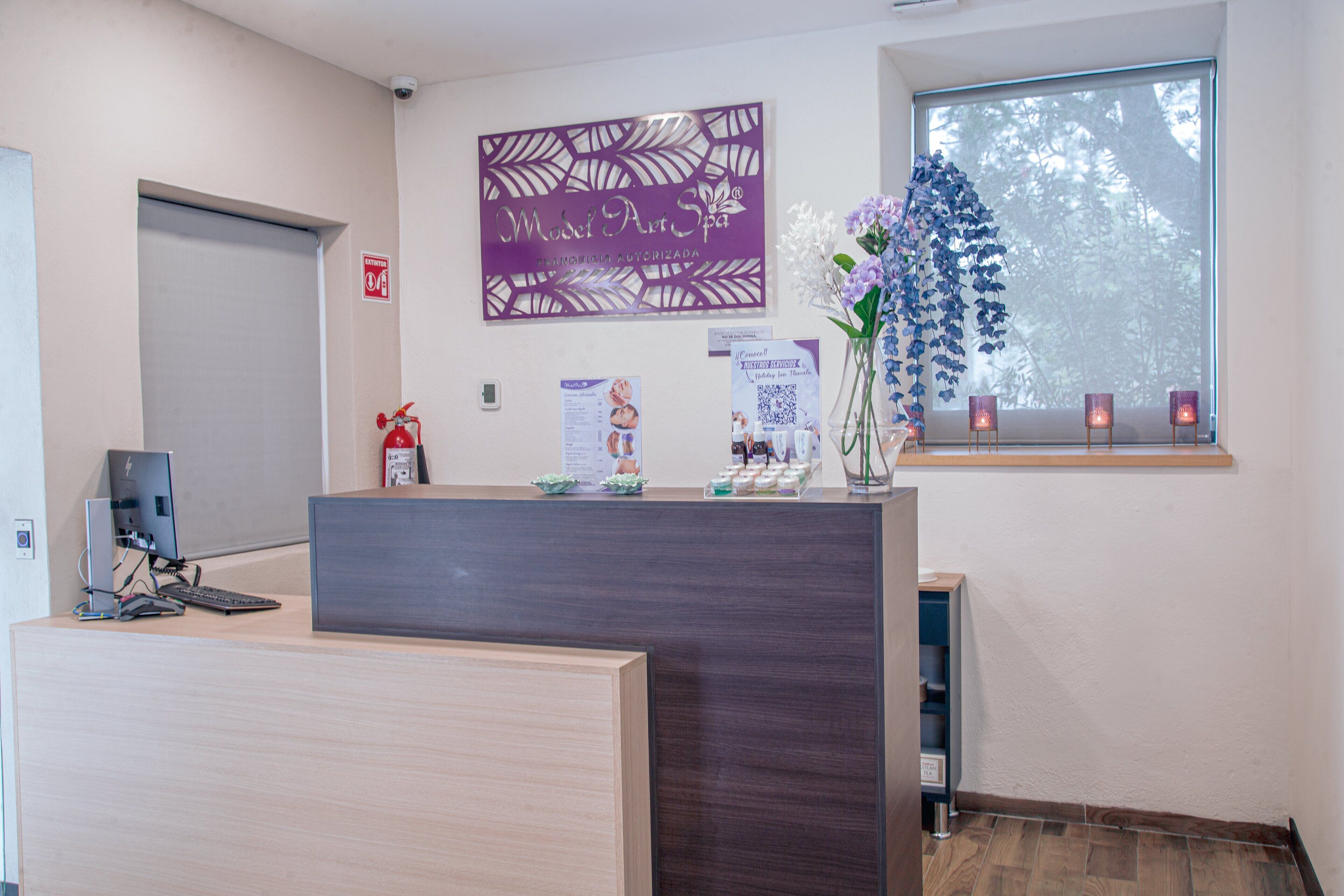 A reception desk with a computer, a fire extinguisher, a decorative sign, a window with a view, and a vase with flowers.