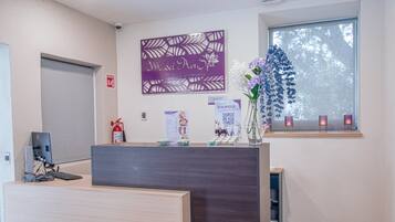 A reception desk with a computer, a fire extinguisher, a decorative sign, a window with a view, and a vase with flowers.