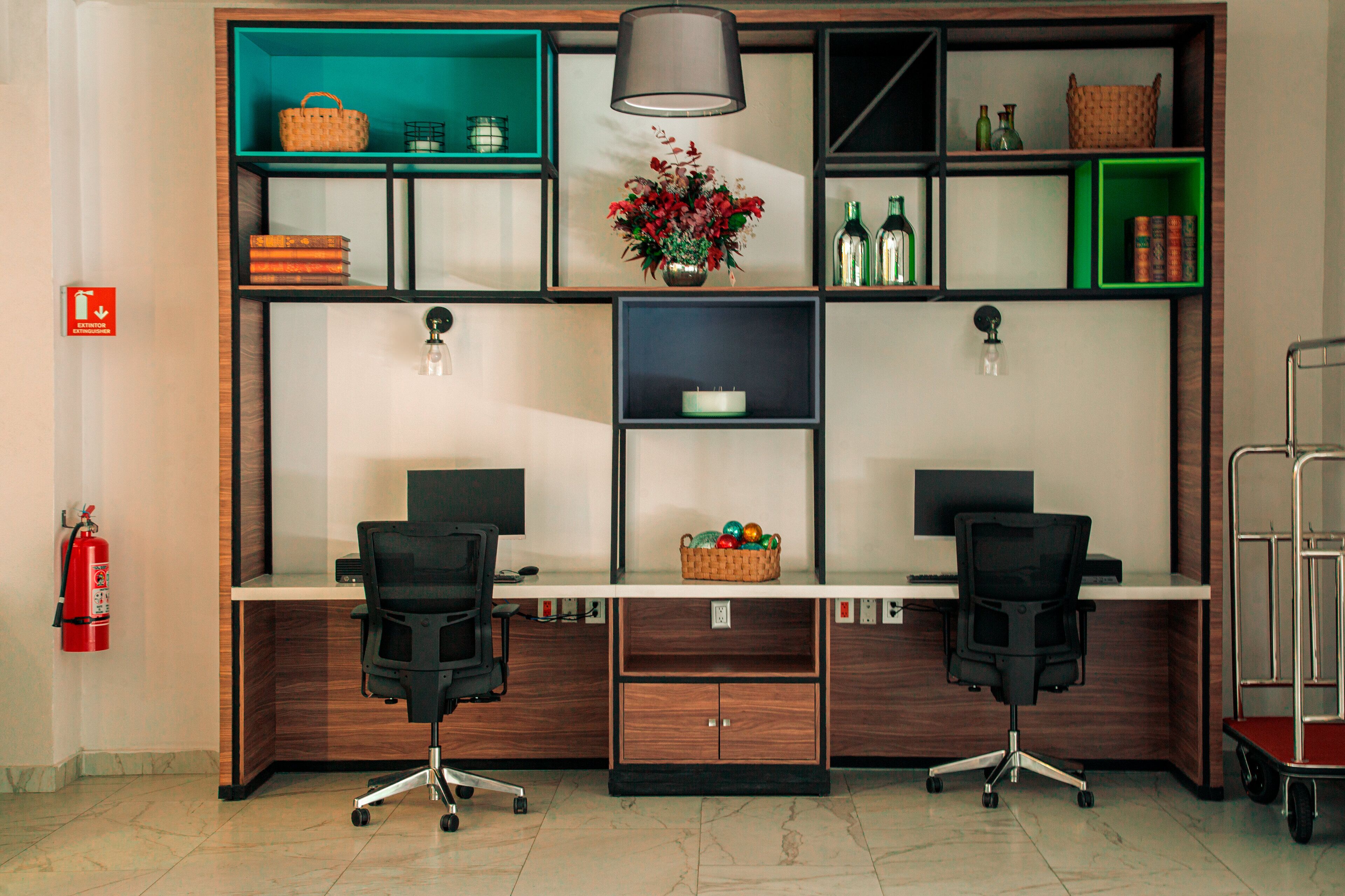 A modern office desk with two black office chairs, a central light fixture, and shelves with various items including a basket, a vase with flowers, and bottles.