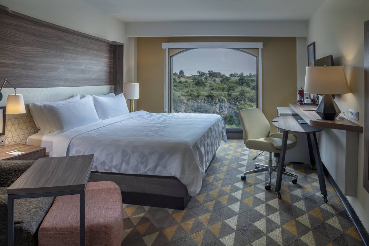 A hotel room with a large bed, a desk with a chair, a lamp, and a view of the outdoors.