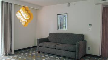 Una habitación con un sofá gris, piso de estampado geométrico, una decoración abstracta grande en la pared, un cuadro enmarcado y una ventana con cortinas.