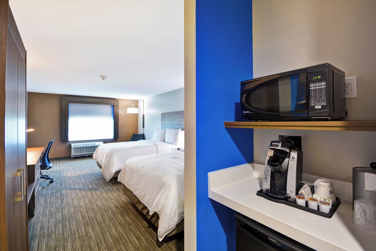 A hotel room with two beds, a desk, a chair, a microwave, a coffee maker, and a window with blinds.