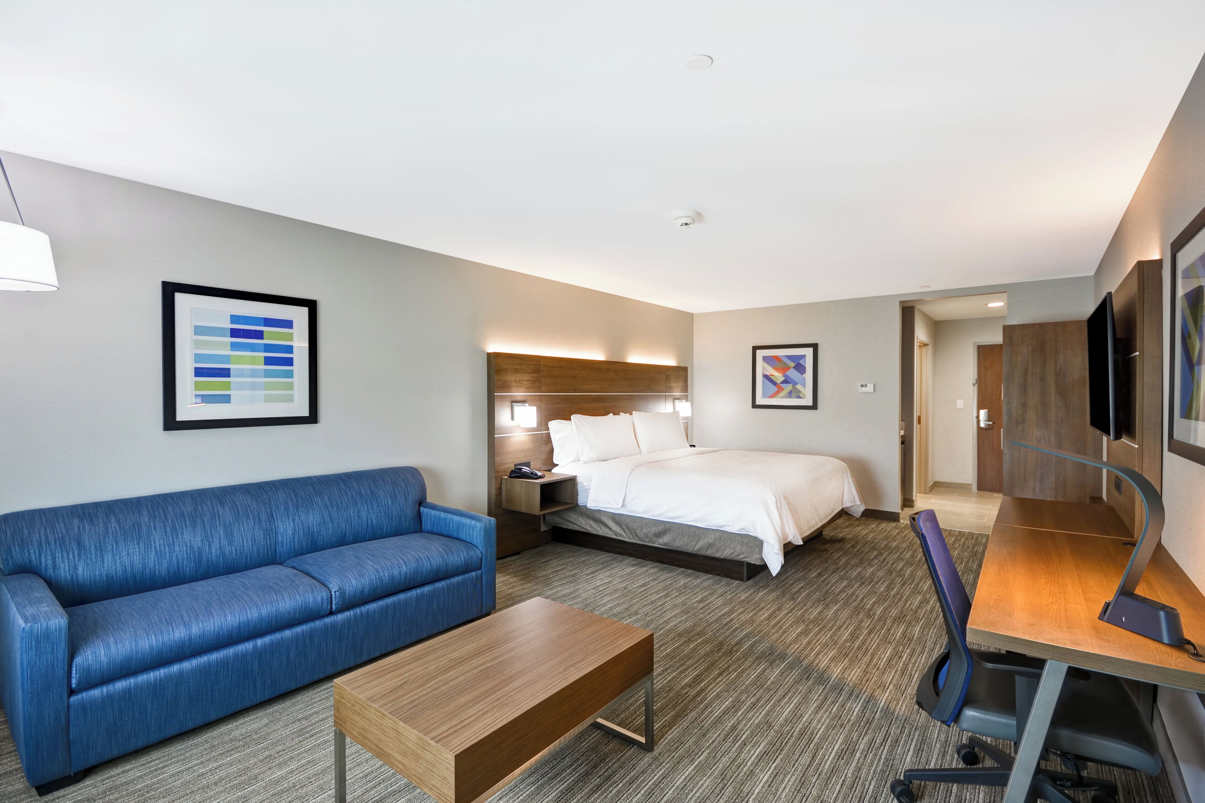 Quarto de hotel com sofá azul, mesa de centro de madeira, cama com roupa de cama branca, escrivaninha com cadeira e televisão na parede.