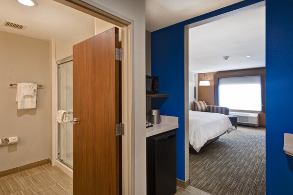 A hotel room with a glass shower door, a small kitchenette, a bed with a white comforter, and a large window providing natural light.