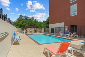 A swimming pool with lounge chairs and a pergola, surrounded by a building with a red facade and a clear sky.