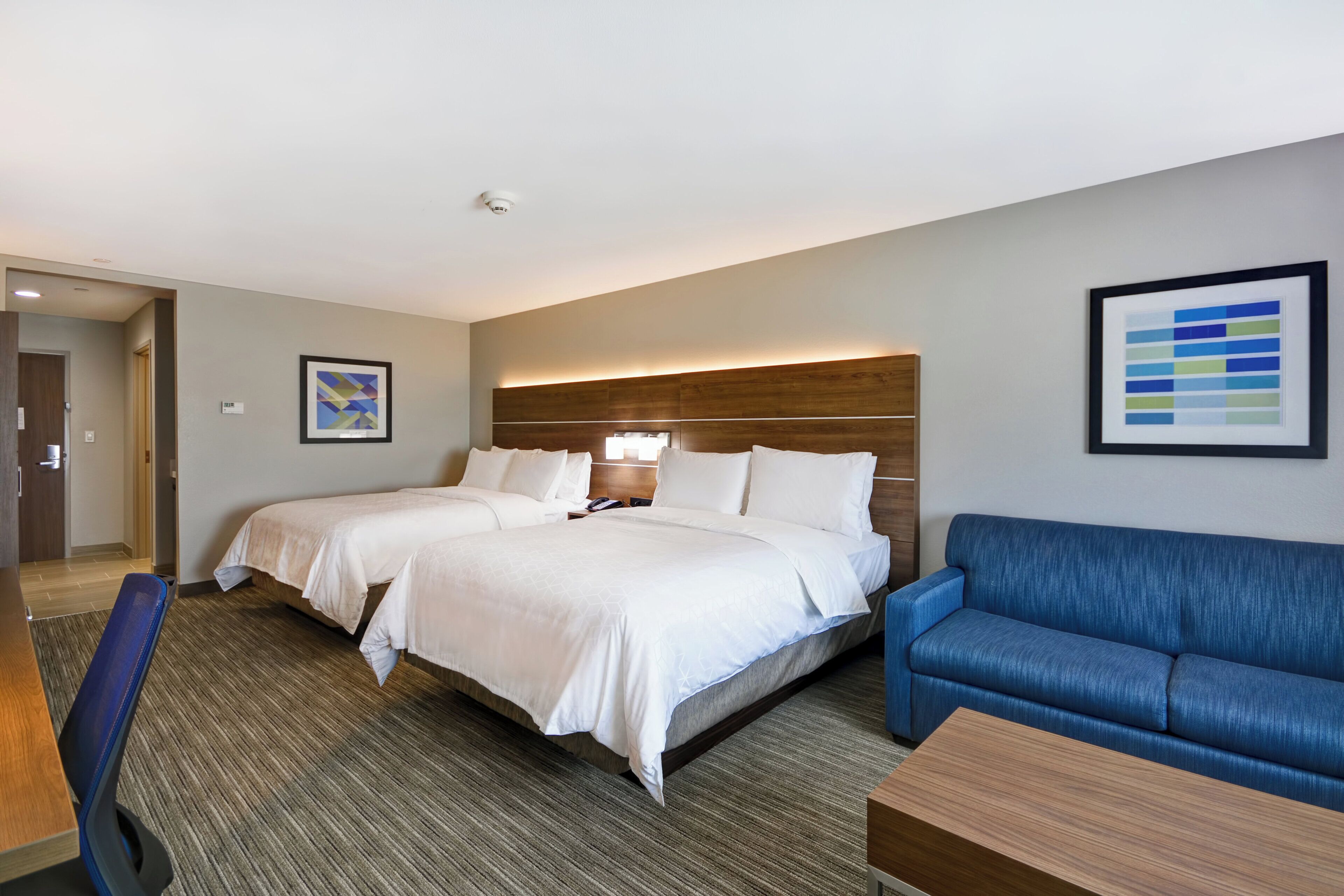 Une chambre d’hôtel avec deux lits, un canapé bleu, un bureau en bois et un tableau abstrait encadré accroché au mur.