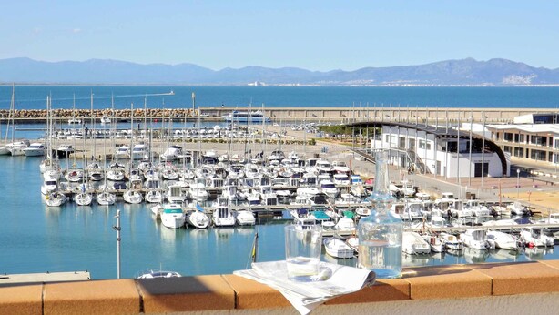 Un port de plaisance où sont amarrés de nombreux bateaux, un pont en arrière-plan et une vue dégagée sur la mer et les montagnes.