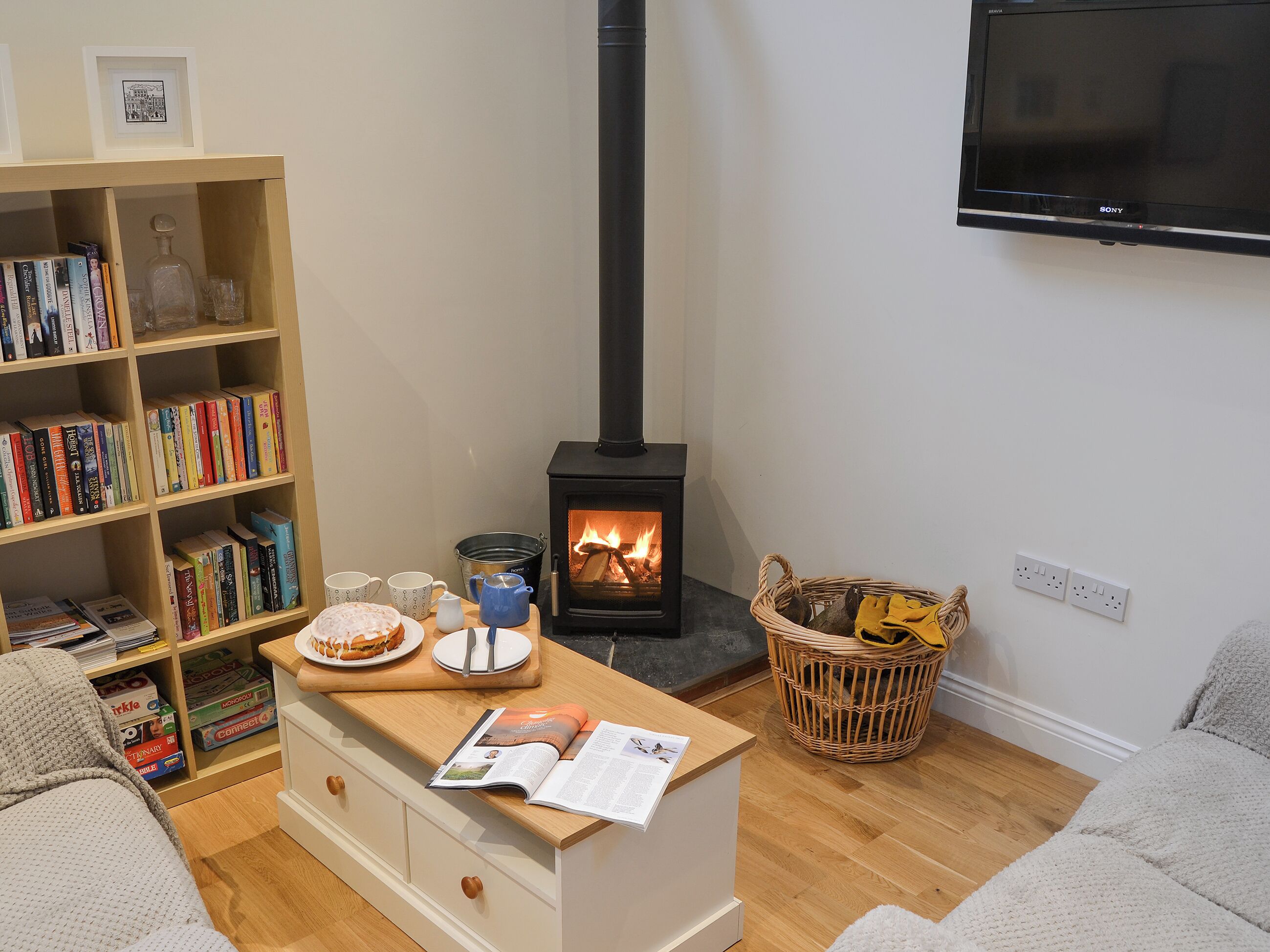 A cozy living room with a bookshelf, a table with a magazine, a fireplace, and a TV mounted on the wall.