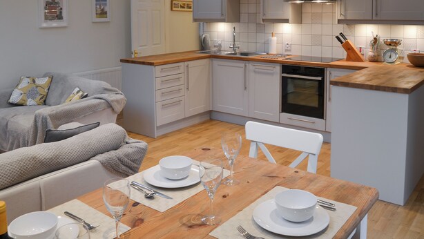 Een moderne keuken met grijze kastjes, een houten aanrechtblad en een eethoek voor vier personen, gedekt met wit servies en wijnglazen.