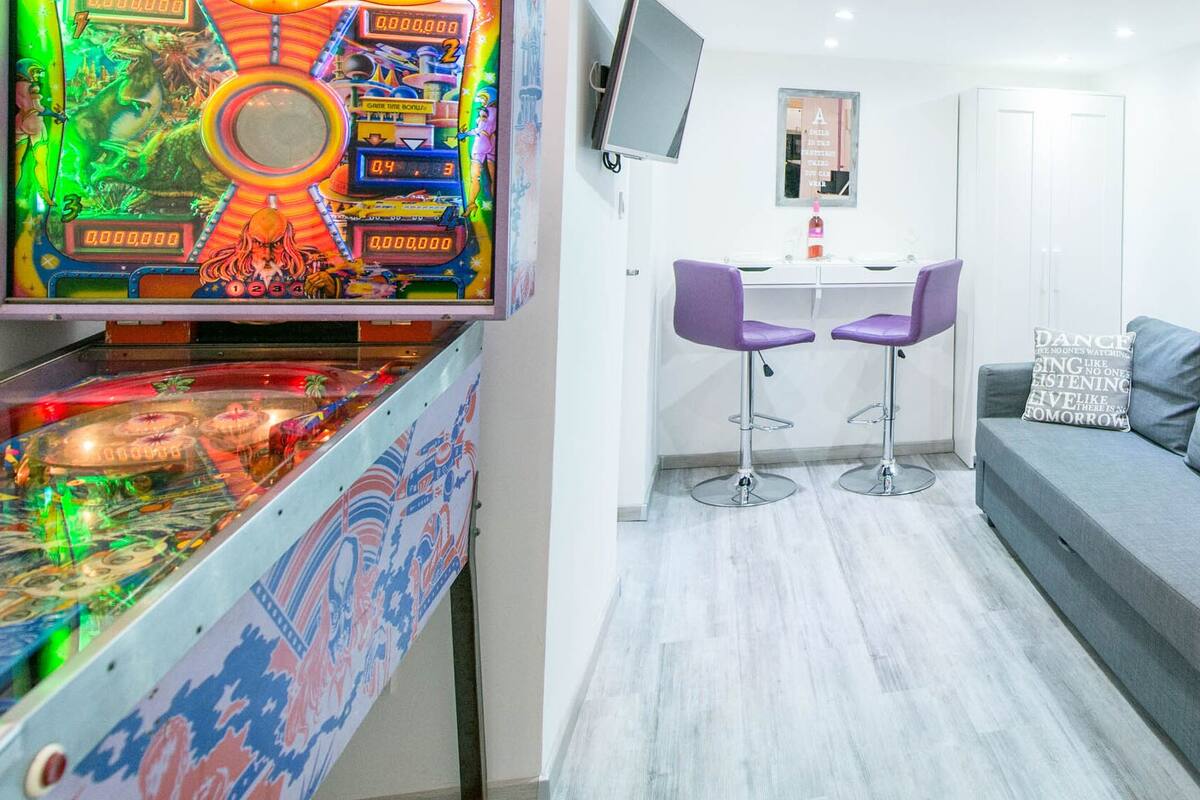 A modern living room with a pinball machine, a grey sofa, a small table with two purple chairs, and a TV mounted on the wall.