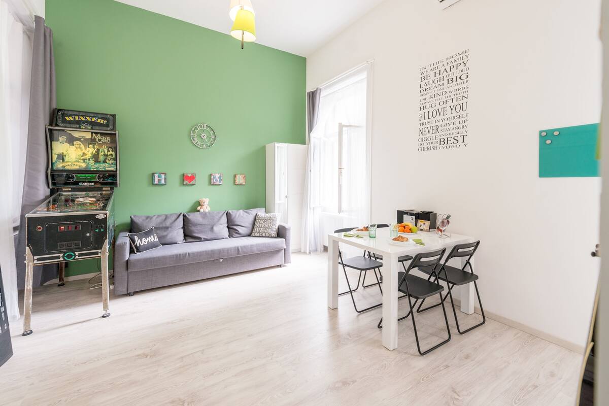 Una sala de estar con una pared verde, un sofá gris, una mesita con sillas, un dardo y una máquina de pinball.