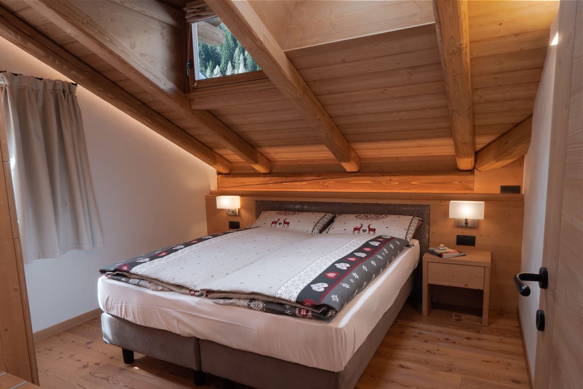 Ein gemütliches Schlafzimmer mit Holzbalkendecke, einem großen Bett mit gemusterten Textilien, zwei Nachttischlampen und Blick auf Bäume durch ein Oberlicht.