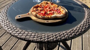 A pizza on a black table with a woven edge, two glasses of white wine, and a bottle of water on a patio with a view of houses and trees. - Olive Tree, Superb gîte, Heated Pool near Carcassonne, overlooking Canal du Midi (Marseillette)