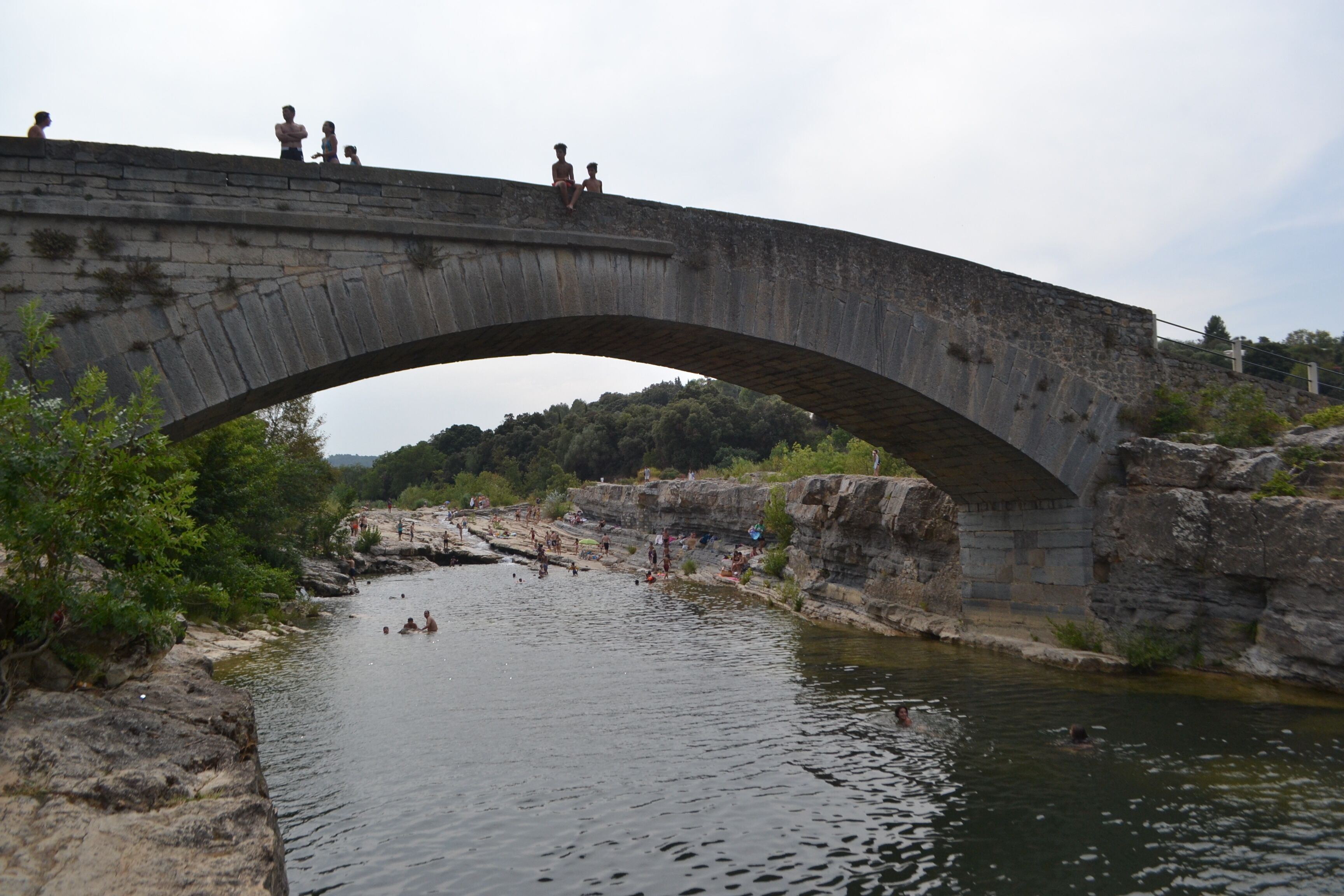 A stone bridge over a river with people on it and swimming in the water below.