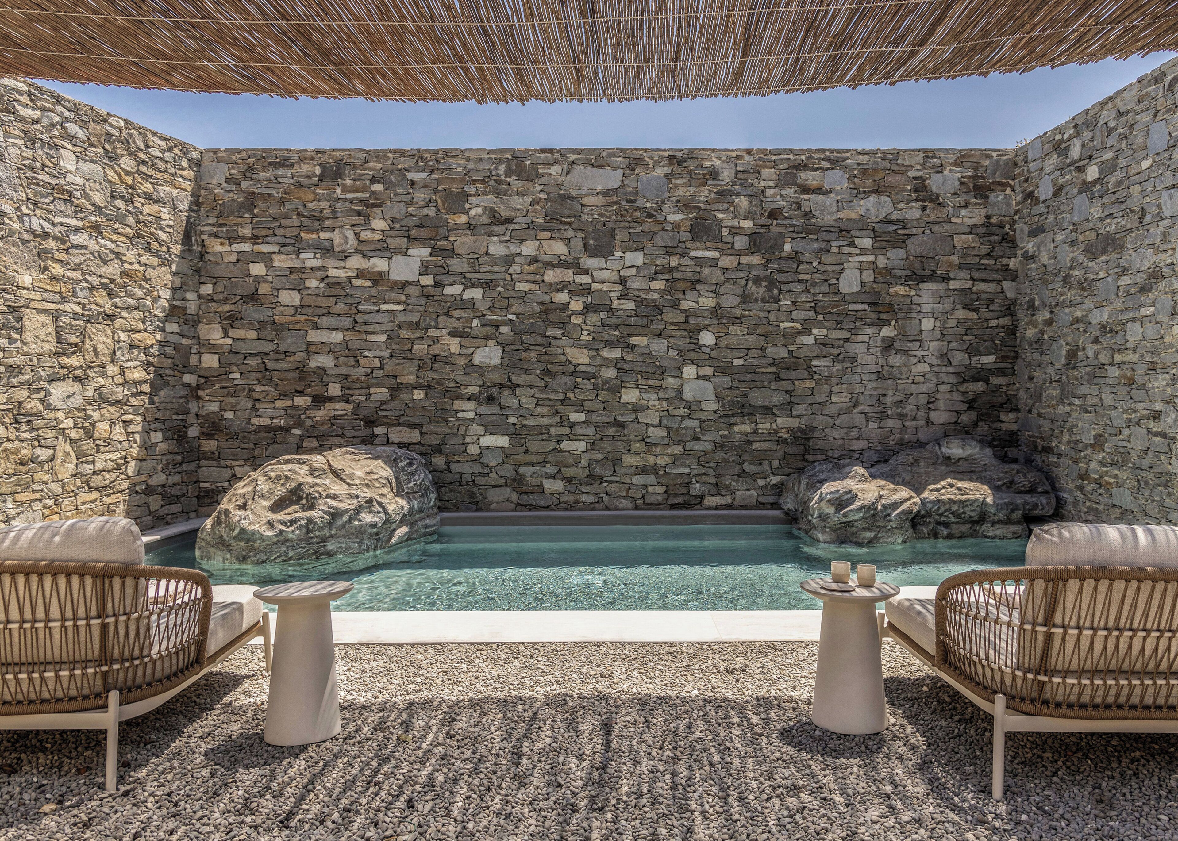 Una piscina infinita de paredes de piedra con dos rocas grandes, rodeada de sillas de mimbre y un piso cubierto de piedras pequeñas.