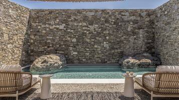 Una piscina infinita de paredes de piedra con dos rocas grandes, rodeada de sillas de mimbre y un piso cubierto de piedras pequeñas.