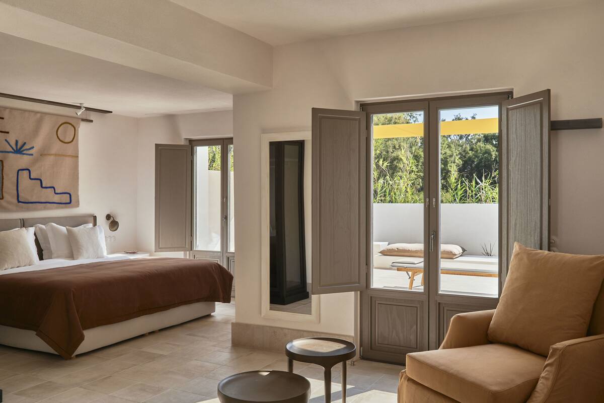 A bedroom with a bed, a chair, a small table, and a large window with an open door leading outside.