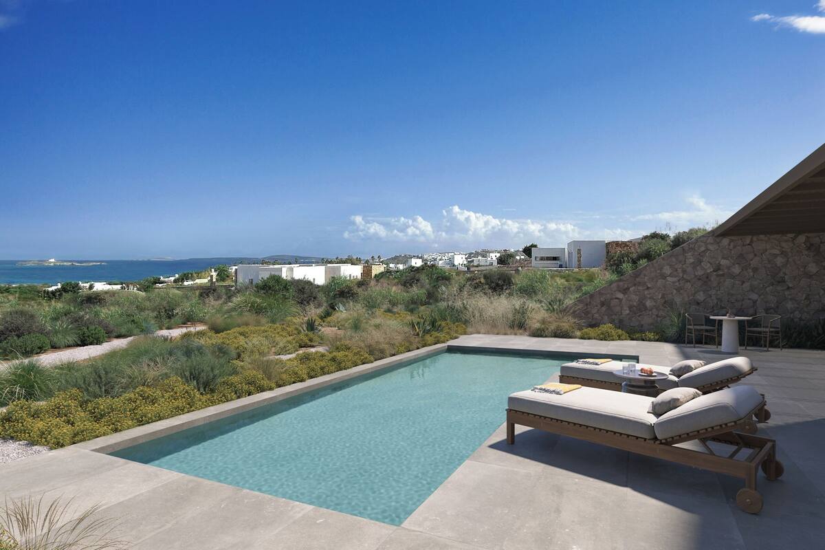 A pool with lounge chairs and a table, surrounded by greenery and a view of the sea.