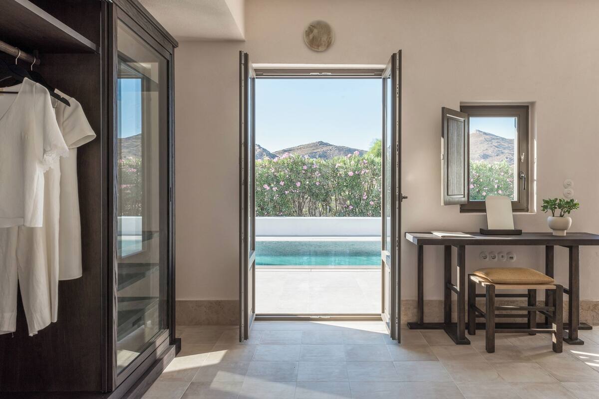 A room with a sliding glass door leading to a pool, a wooden chair, a small table with a mirror, and a cabinet with hanging clothes.